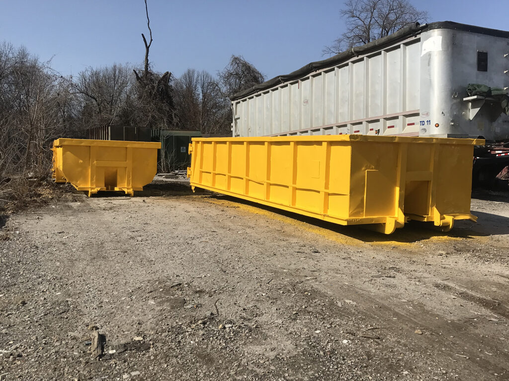 Two large yellow dumpsters are placed on a gravel area near a white industrial truck in an outdoor setting.