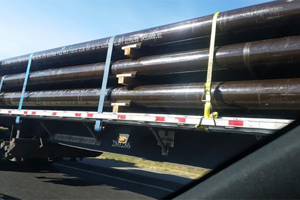 Large metal pipes secured with straps are being transported on a flatbed trailer on a highway.