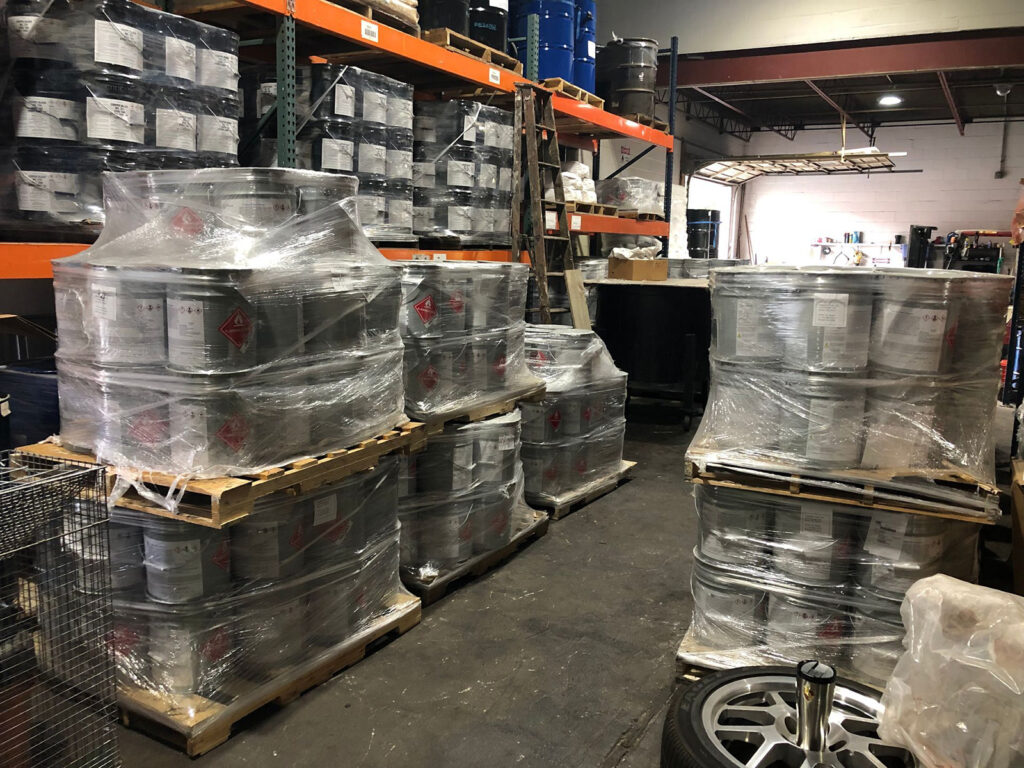 Warehouse with multiple pallets stacked with metal drums wrapped in plastic, surrounded by shelves of similar containers.