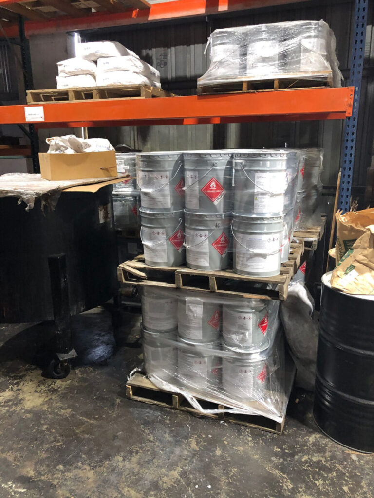 Metal containers with hazard labels are stacked on pallets in a warehouse. Shelves hold additional wrapped packages.