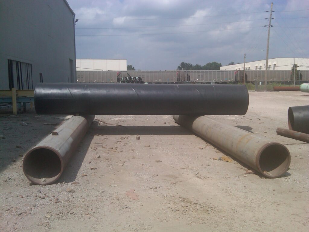 Large black pipe supported by two rusty pipes in an industrial outdoor setting with buildings in the background.