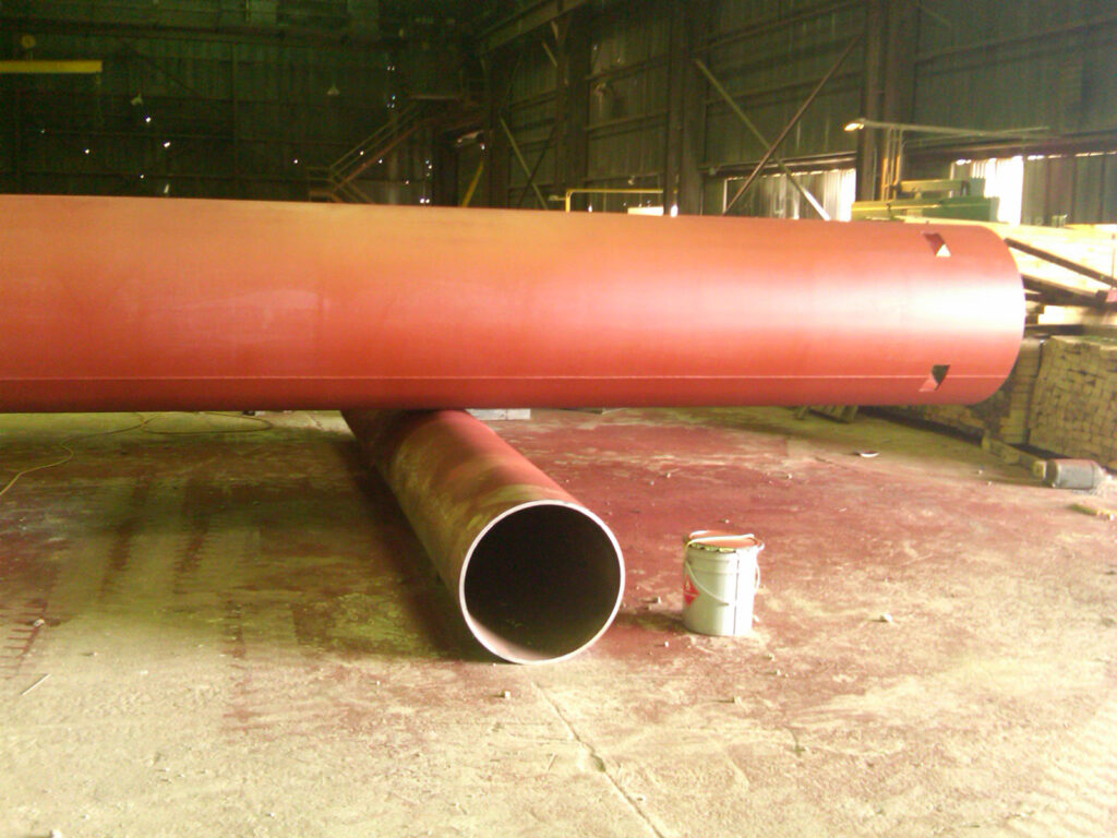 Large red cylindrical metal pipe resting on smaller pipe in an industrial warehouse. A paint bucket is nearby.